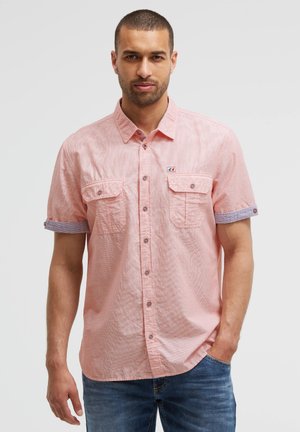 Mann, der ein hellrosa kurzärmeliges Hemd mit zwei Brusttaschen und blauen hochgekrempelten Ärmeln trägt, kombiniert mit blauen Jeans.