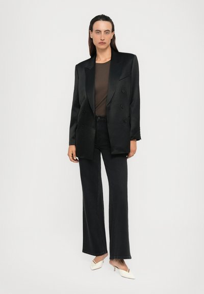 Persoon in zwarte dubbelgeknoopte blazer, bruine top, zwarte wijde jeans en witte puntige hakken, staande tegen een effen achtergrond.