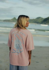 Różowa, oversize'owa koszulka z niebieską grafiką lwa i fali, z napisem "SURF IN THE BRAND WITH THE WAVE" na plecach.