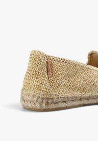 Flettet strå-sutsko med rund tå, beige farve, tekstureret overflade og naturlig rebdetaljering langs sålen. Diskret logo-tag vedhæftet.