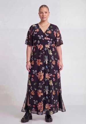 Kvinde stående iført en mørk maxikjole med farverigt blomsterprint, korte flagrende ærmer, sorte kraftige støvler, guldsmykker og en neutral baggrund.