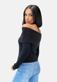 Schwarzer, schulterfreier, gerippter Strickpullover mit langen Ärmeln; figurbetontes Design und strukturierte Details; kombiniert mit hellblauen Jeans.