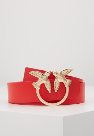 Ceinture en cuir rouge avec une boucle circulaire dorée ornée de deux oiseaux détaillés se faisant face sur une surface blanche.