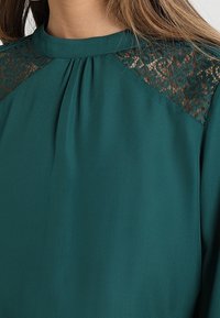 Tealblus med hög hals, som har spetsdetaljer på axlarna och plissering fram. Slät tyg och avslappnad passform.