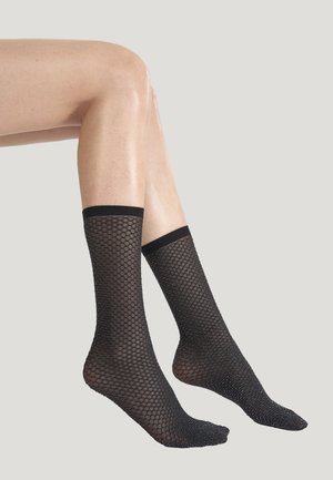 Ben med svarta knähöga nätstrumpor med diamantmönster mot en enfärgad ljusgrå bakgrund.