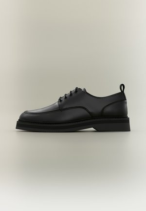 Chaussure habillée en cuir noir à lacets, semelle épaisse et languette de traction au talon, montrée de profil sur un fond neutre.