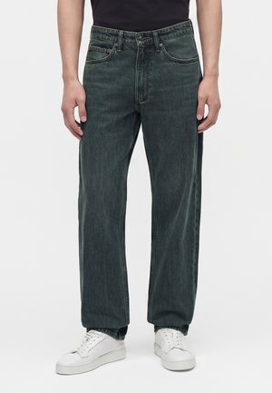 Homme portant un jean droit vert foncé et des baskets blanches, debout devant un fond clair uni.