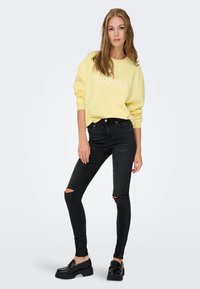 Sweatshirt galben deschis cu text alb "SUPERB", asortat cu blugi negri skinny cu rupturi și mocasini negri; potrivire lejeră, textură moale.