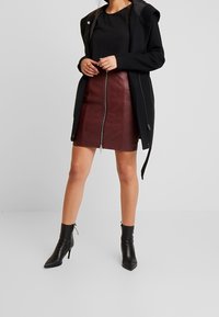 Giacca nera con cappuccio sopra una maglia nera testurizzata, abbinata a una gonna in pelle bordeaux con zip e stivaletti neri con punta affilata.