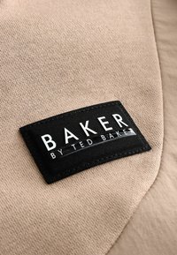 Béžová tkanina s čiernou textilnou etiketou, na ktorej je biele písmo "BAKER" a "by TED BAKER". Etiketa má prešité okraje.