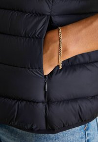 Czarna kurtka puchowa o pikowanym wzorze, z zamykaną kieszenią boczną. Widoczna złota bransoleta na nadgarstku, poniżej materiał dżinsowy.