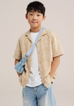 Garçon portant une chemise beige à fleurs à manches courtes, un t-shirt blanc, un jean bleu clair et un sac bandoulière bleu, debout les mains dans les poches.