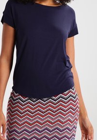Granatowa bluzka z krótkim rękawem o okrągłym dekolcie, zestawiona z wielokolorową spódnicą w zygzakowy wzór z przewagą czerwonego, białego i czarnego.