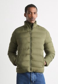Olivengrønt puffjakke med høj krave, lynlåslukning og lodret syning. Båret over en mørk langærmet top og blå jeans.