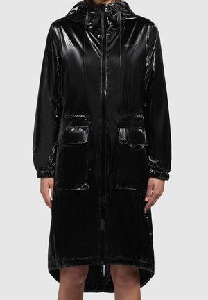 Žena v lesklém černém pláštěnkovém kabátu s kapucí, předním zipem a velkými předními kapsami, stojící před jednoduchým šedým pozadím.