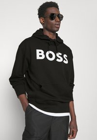 Felpa nera in misto cotone, con un grande logo bianco "BOSS" sul davanti, dotata di tasca a marsupio e cappuccio regolabile con cordino.