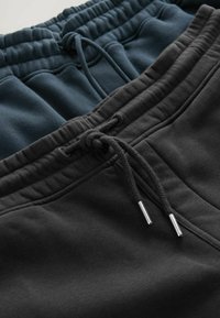 Dos pares de pantalones de chándal: uno en un color verde azulado oscuro y otro negro, que presentan cinturillas elásticas con cordones ajustables y puntas metálicas plateadas.