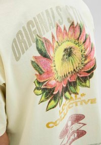Krēmkrāsas tienīte ar lielu grafiku, kurā attēlota krāsaina, ziedoša zieds, un uzraksti "ORIGINAL'S STORE" un "LOCAL COLLECTIVE."