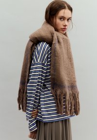 Eșarfă maro din lână cu capete franjurate, așezată peste o cămașă cu mâneci lungi în dungi albastre și albe, cu croială lejeră și buzunar lateral.