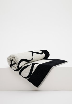 Mustavalkoinen kudottu kangas, jossa on abstrakteja kaarevia viivoja, osittain rullattu ja valkoisen pinnan päällä roikkuva.