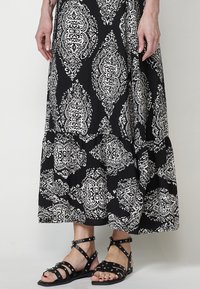 Jupe maxi noire avec un motif orné blanc, présentant un bas en plusieurs niveaux. Associée à des sandales plates noires, agrémentées de petits clous.