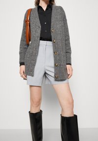Grijze gebreide cardigan met een knoopsluiting aan de voorkant, gecombineerd met een zwarte button-up shirt en lichtgrijze geplooide shorts. Zwarte knielange laarzen.
