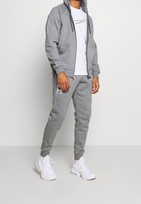 Sudadera deportiva gris con cremallera y bolsillos, combinada con pantalones de chándal grises a juego con un logo. Completado con zapatillas deportivas blancas.