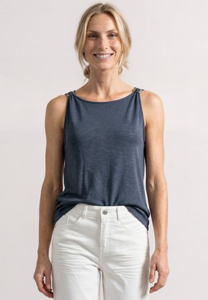 Femme souriante aux cheveux blonds portant un haut bleu foncé sans manches et un pantalon blanc, debout devant un fond uni et clair.