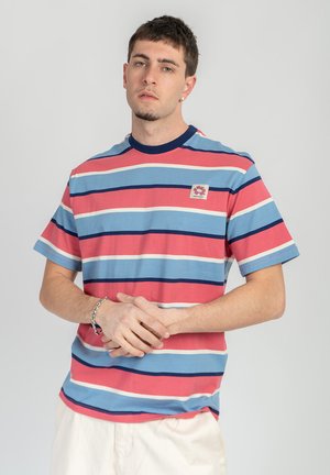 Young man wearing a short-sleeve striped t-shirt in blue, red, white, and navy, with light-colored pants and a silver bracelet, standing against a plain background.