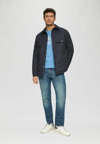 Navy button-up jacket with two chest pockets, worn over a light blue graphic t-shirt, paired with light wash jeans and white sneakers.
