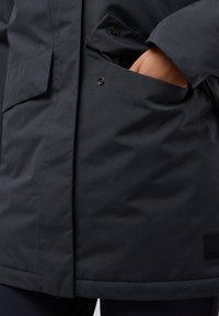 Donkerblauwe jas met een gladde, matte textuur. Voorzien van een zijzak met zwarte drukknoopsluiting en een merkje nabij de zoom.