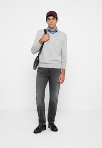Suéter cinza em V, camisa azul de colarinho, jeans cinza e mocassins pretos, finalizado com um boné bordô e uma mala de ombro preta.