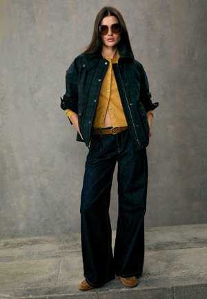 Czarna kraciasta kurtka z futrzanym kołnierzem, żółty sweter zapinany na guziki, ciemne dzwony i brązowe buty. Duże ciemne okulary przeciwsłoneczne dopełniają wygląd.