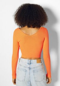 Jasnopomarańczowy prążkowany crop top z długim rękawem, z głębokim dekoltem z tyłu. W połączeniu z jasnoniebieskimi dżinsami z wysokim stanem.