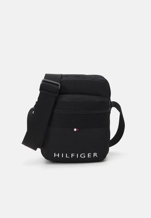 Kompaktowa czarna torba na ramię z regulowanym paskiem, przednią kieszenią na zamek, małym logo flagi i napisem „HILFIGER” na dole na środku.