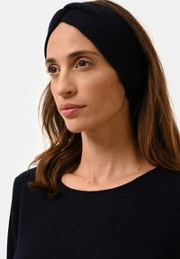 Černý pletený čelenka se zkrouceným designem, měkkou texturou a těsným střihem, v kombinaci s tmavou svetrem s širokým výstřihem.