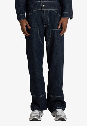 Jeans denim blu scuro con cuciture bianche a contrasto, tasche frontali applicate, indossati con sneakers argento e bianchi.