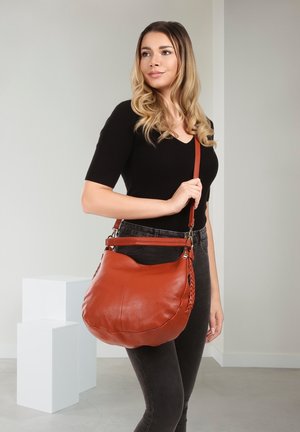 Femme aux cheveux blonds ondulés portant un haut noir et un jean foncé, tenant un grand sac à bandoulière en cuir marron avec un détail tressé.