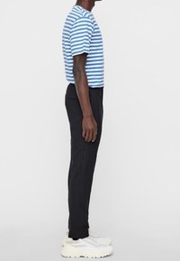 Muž stojící bokem, oblečený v modro-bílém pruhovaném triku s krátkým rukávem, černých kalhotách a b�ílých chunky teniskách na jednobarevném pozadí.