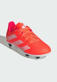 Punainen jalkapallosandaali, jossa on teksturoitu pinta, kirkkaan vaaleanpunaiset yksityiskohdat, valkoinen logo ja kuusi mustaa kumikärkeä pohjassa pidon parantamiseksi.