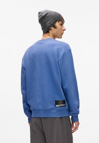 Modrá mikina s žebrovanými manžetami a lemem, s černým štítkem s bílým textem. Jednotlivec nosí šedou čepici.