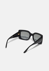 Óculos de sol quadrados pretos com lentes cinzentas, apresentando armações grossas e um design texturizado nas hastes. Logotipo dourado na parte interna do braço esquerdo.