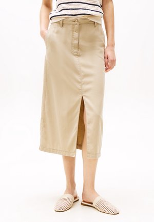 Kvinde iført beige midinederdel med frontslids, stribet ærmeløs top og vævede beige slip-on sko, stående med hænderne i lommerne.