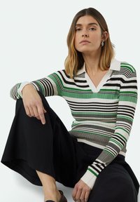 Gestreifter grüner und schwarzer Strickpullover mit V-Ausschnitt und weißem Kragen, kombiniert mit einem schwarzen Rock. Modell sitzt und zeigt die Details der Ärmel.