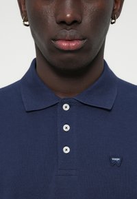 Σκούρο μπλε πόλο μπλουζάκι με υφαντή υφή, τρεις κουμπιά στη μανσέτα, και ένα μικρό κεντημένο λογότυπο "Wrangler" στη αριστερή πλευρά.