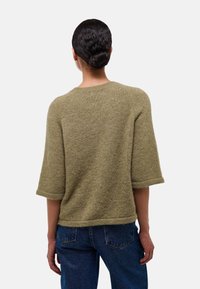 Femme aux cheveux bruns en chignon, portant un pull en tricot olive ample avec des manches jusqu'aux coudes et un jean bleu, de dos.