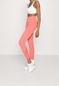Różowe legginsy wysokiej talii z elastycznego materiału mają gładką teksturę oraz kontrastowe białe logo Nike blisko kostki. Noszone z białym stanikiem sportowym.