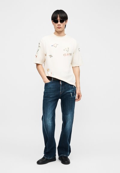 Άνδρας που φοράει μαύρα γυαλιά ηλίου, λευκό υπερμεγέθες T-shirt με γραφικά σχέδια, μπλε τζιν και μαύρα παπούτσια, στέκεται με το ένα χέρι στην τσέπη.