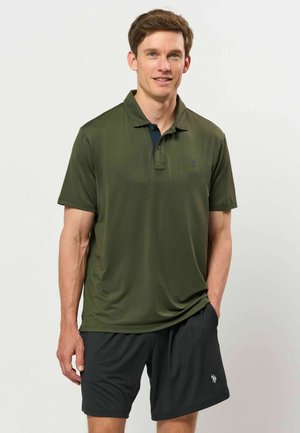 Jeune homme portant un polo à manches courtes vert olive et un short noir, debout avec une main dans la poche devant un fond uni.