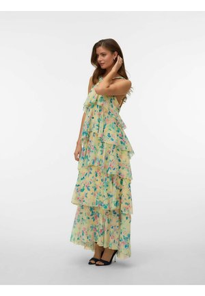 Frau trägt ein ärmelloses, gelbes, geblümtes, mehrlagiges Maxikleid und schwarze offene High Heels, posiert mit einer Hand in den Haaren.